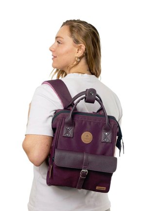Young woman wearing white shirt carries a purple leather and fabric backpack with a front buckle pocket, facing sideways against a white background.
