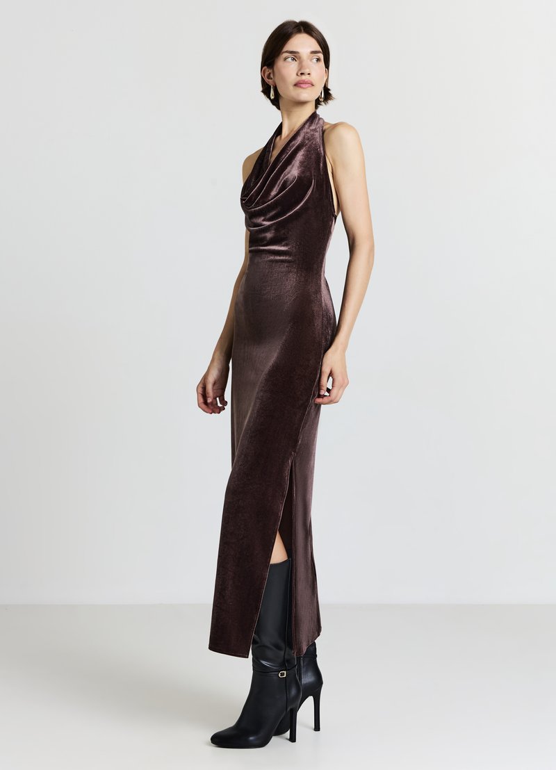 Abito in velluto con collo all'americana di colore marrone scuro, con davanti drappeggiato, spacco laterale e vestibilità aderente, abbinato a stivali neri con tacco alto.