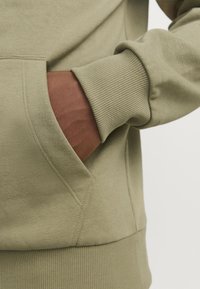 Olijfgroen sweatshirt met een kangoeroezak, geribbelde manchetten en een soepele textuur. Close-up van de zak met een hand erin.