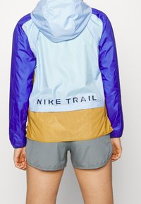 Kurtka Trail Nike w niebiesko-fioletowym górnym kawałku, z dolną częścią w kolorze złotym. Lekki, wodoodporny materiał z kapturem i logo.