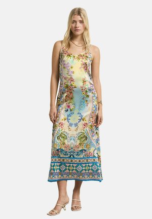 Woman standing, wearing a sleeveless, ankle-length dress with colorful floral and geometric patterns, and open-toed heeled sandals.