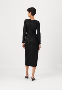 Vestido preto de mangas longas com textura floral; silhueta ajustada, comprimento midi, tecido suave e fenda nas costas. Usado com sandálias de salto alto pretas.