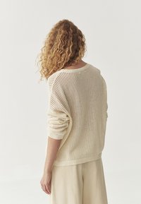 Leichter, krämiger, gestrickter Pullover mit lockerem Schnitt, breitem Halsausschnitt und hochgerollten Ärmeln, vor einem schlichten Hintergrund.