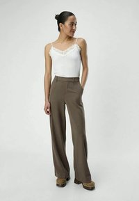 Camisola bianca con pizzo, abbinata a pantaloni marroni a vita alta e a gamba larga, che mostrano una vestibilità su misura e una texture di tessuto liscia.