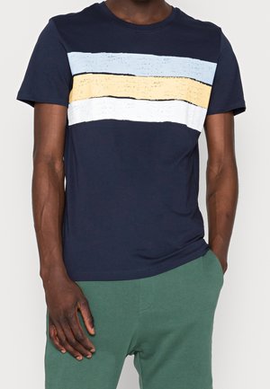 Mann trägt ein marineblaues T-Shirt mit kurzen Ärmeln, das hellblaue, gelbe und weiße horizontale Streifen über der Brust aufweist, kombiniert mit grünen Hosen.