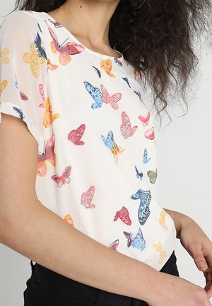 Valkoiseen lyhythihaiseen puseroon painettu värikkäitä perhoskuvioita, jossa kiharat hiukset omaava henkilö, yhdistettynä mustiin housuihin.