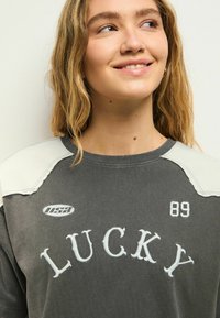 Sweatshirt gris avec "LUCKY" en broderie blanche, accents d'épaule gris clair, et col ras du cou décontracté avec le détail du numéro "89".