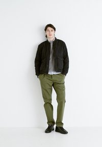 Chaqueta bomber acolchada negra sobre un suéter gris, con pantalones verdes y zapatillas negras. Diseño minimalista con un ajuste casual y un look en capas.