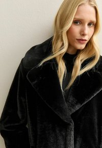 Blondynka w dużym czarnym futerku z materiału syntetycznego na tle jasnej, gładkiej ściany, z luźnymi falami opadającymi na jej twarz.