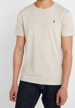 Man wearing a beige short-sleeve T-shirt with a small brown embroidered logo on the left chest and dark blue jeans.