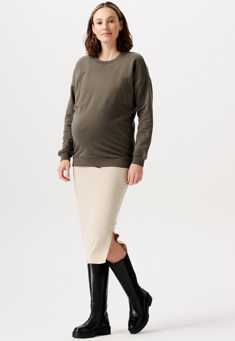 Schwangerschafts-Sweatshirt in Olivgrün, gerippter cremefarbener Rock mit Seitenschlitzen und knielangen schwarzen Stiefeln mit strukturierten Sohlen, stehend.