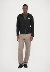 Man wearing black zip-up hoodie with EA7 logo, olive green shirt, beige textured pants, and black leather shoes standing against white background.