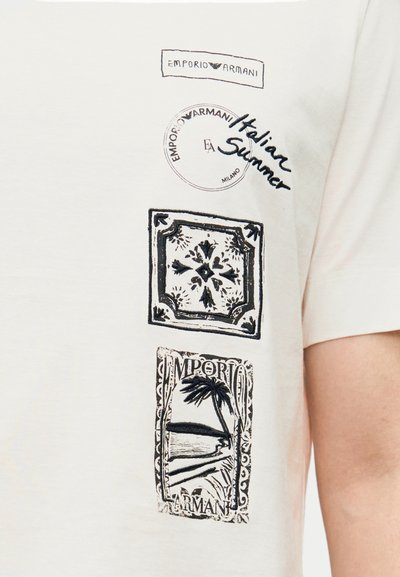 Λευκό βαμβακερό μπλουζάκι με μαύρα τυπωμένα σχέδια, που περιλαμβάνουν σφραγίδες με το λογότυπο "Emporio Armani" και τροπικά μοτίβα, δημιουργώντας μια υφασμάτινη όψη.