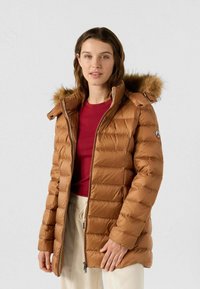 Giacca a piumino marrone con cuciture orizzontali, cappuccio con bordo in pelliccia, chiusura frontale con cerniera e tasche laterali. Indossata sopra una maglia rossa.