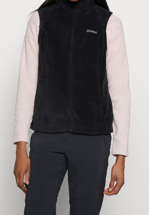 Persoon draagt een zwarte Columbia fleece bodywarmer met rits over een lichtroze top met lange mouwen en donkere broek, staand tegen een effen achtergrond.