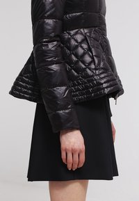 Quiltet sort dunjakke med en blank finish, tætsiddende talje, udvidet kant og lange ærmer. Lynlåslomme ved taljen.