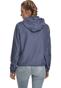 Giacca con cappuccio di colore blu navy realizzata in materiale leggero, dotata di polsini elastici e vestibilità comoda, con una texture liscia e lucida.
