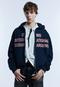 Marineblauer Kapuzenpullover mit durchgehendem Reißverschluss und großem roten Text "INTERNATIONAL ARCHIVES" auf der Vorderseite. Wird über einem weißen Shirt und dunklen Jeans getragen, mit Taschen ausgestattet.