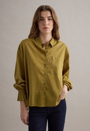 Joven mujer con cabello ondulado castaño, vestida con una camisa de manga larga verde oliva abotonada y pantalones oscuros, parada con una mano en el bolsillo.