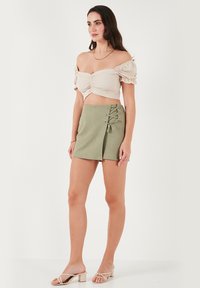 Beige off-shoulder cropped top met gathered stof, gecombineerd met hoge groene shorts met een vetersluiting aan de zijkant.