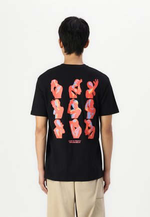 Homme portant un t-shirt noir avec neuf figures humaines abstraites rouges, bleues et roses en différentes poses au dos, debout devant un fond blanc.