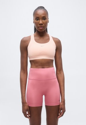 Mujer atlética que lleva un sujetador deportivo melocotón y unos pantalones de ciclista rosa de cintura alta, de pie contra un fondo claro y sencillo.