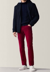 Marine peacoat over een kabelgebreide trui, gecombineerd met rode corduroy broek en witte sneakers. De outfit heeft een textuurdesign en klassieke vormen.