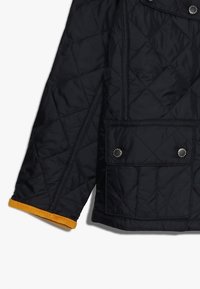 Gestepptes schwarzes Jackett mit einem gelben Rippbündchen, zwei Fronttaschen und Druckknöpfen. Die Textur ist glatt und das Design ist strukturiert.