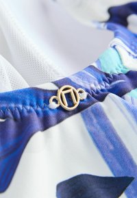 Detaliu al unui top de costum de baie, prezentând o țesătură albă texturată, un model abstract în nuanțe de albastru și aqua, și un închizător din metal aurit.