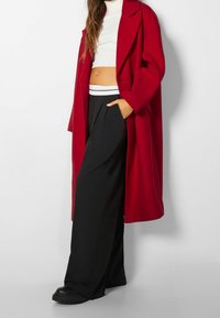 Abrigo largo rojo con cuello de muesca, usado sobre un top blanco corto y pantalones anchos negros. Botas negras con suela gruesa completan el atuendo.