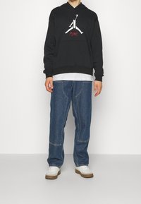 Felpa nera con cappuccio e logo Jumpman bianco, abbinata a jeans blu larghi con dettagli di cucitura e sneakers bianche.