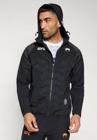 Veste noire avec un motif géométrique texturé, fermeture éclair frontale, capuche ajustable et accents de logo. Le matériau semble léger et sportif.