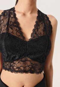 Bralette negro de encaje con patrones florales en el torso de una mujer, con paneles transparentes y bordes festoneados.