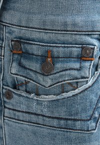 Niebieska tkanina dżinsowa ma małą kieszeń z pomarańczowymi ściegami i metalowymi elementami. Zniszczona tekstura z widocznym wzorem zużycia.