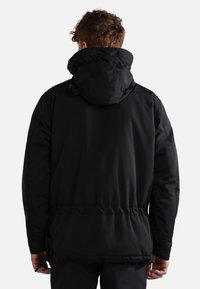 Homme aux cheveux bouclés portant une veste noire à capuche, vu de dos sur un fond clair uni.