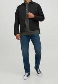 Svart knappjacka över en grön T-shirt, i kombination med blå jeans och marinblå sneakers. Texturerat tyg, avslappnad passform och synliga fickdetaljer.