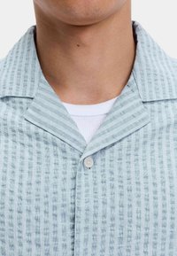 Close-up van de nek en bovenste borst van een persoon die een lichtblauwe geblokte overhemd met knopen draagt over een wit ribbelshirt.