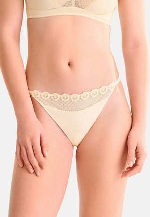 Femme portant un ensemble de lingerie en dentelle beige, mettant l'accent sur la taille et les hanches, avec des détails en dentelle florale sur la culotte et la bande du soutien-gorge.