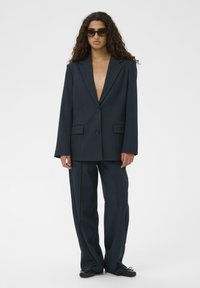 Tengerészkék, túlméretezett blézer bevágott hajtókkal, egysoros gombolással és oldalzsebekkel, széles szárú tengerészkék nadrággal párosítva. Sima textúra.