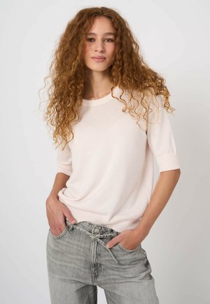 Młoda kobieta z długimi kręconymi rudymi włosami, ubrana w jasnoróżową bluzkę z krótkim rękawem i wysokim stanem jasnoszarych dżinsów, z rękami w kieszeniach.