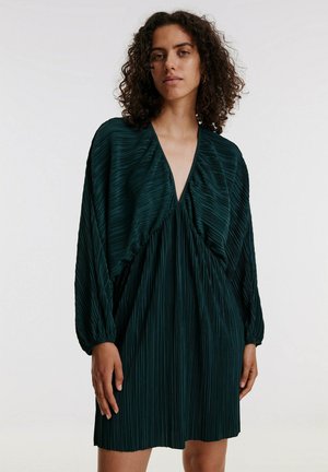 Dunkelgrünes, plissiertes Kleid mit tiefem V-Ausschnitt und langen, puffigen Ärmeln. Der Stoff hat ein strukturiertes, geripptes Muster. Midi-Länge bis zur Mitte des Oberschenkels.