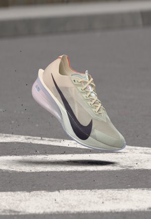 Svetlo béžová bežecká topánka Nike s čiernym logom vo vzduchu nad sivou cestou označenou bielymi čiarami.