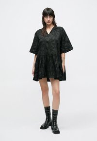 Μαύρο φόρεμα με κοντά μανίκια και V-λαιμόκοψη, υφασμένο με υφή, με φαρδιά σιλουέτα και σούρες στη μέση. Συνδυάζεται με μαύρες μπότες μέχρι το γόνατο.