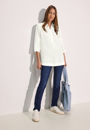 Blouse en coton blanc avec un col et des manches trois-quarts, associée à un jean bleu foncé et des baskets blanches ; sac fourre-tout en denim bleu tenu à la main.