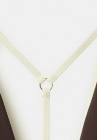 Cinghie di tessuto crema con cuciture a zigzag collegate da un piccolo anello metallico argentato su sfondo bianco.