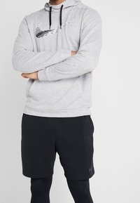 Homme portant un sweat à capuche gris clair avec un logo graphique noir, un short de sport noir et des collants noirs, bras croisés sur un fond blanc uni.