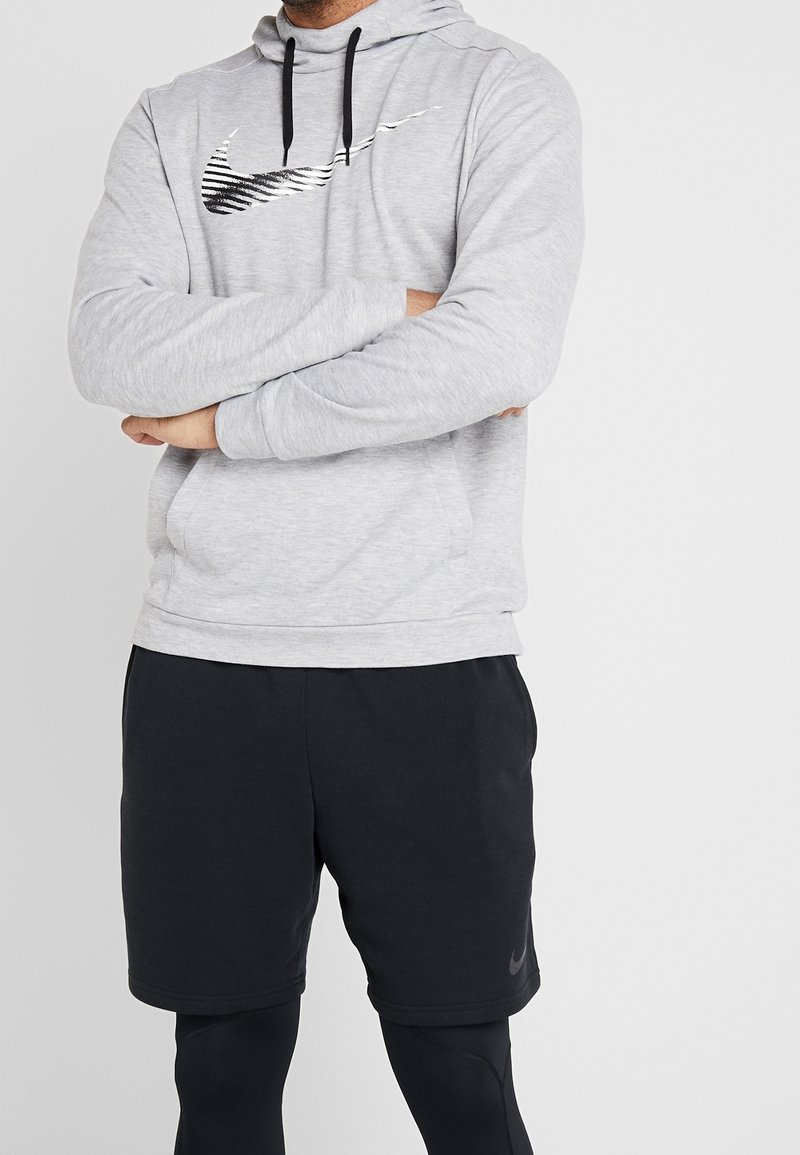 Homme portant un sweat à capuche gris clair avec un logo graphique noir, un short de sport noir et des collants noirs, bras croisés sur un fond blanc uni.