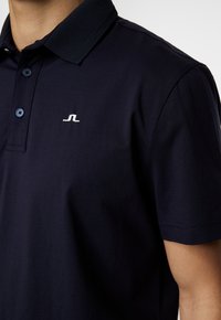 Mörkblå pikétröja i bomull med en texturerad krage, tre knappar och en liten vit logotyp på bröstet.