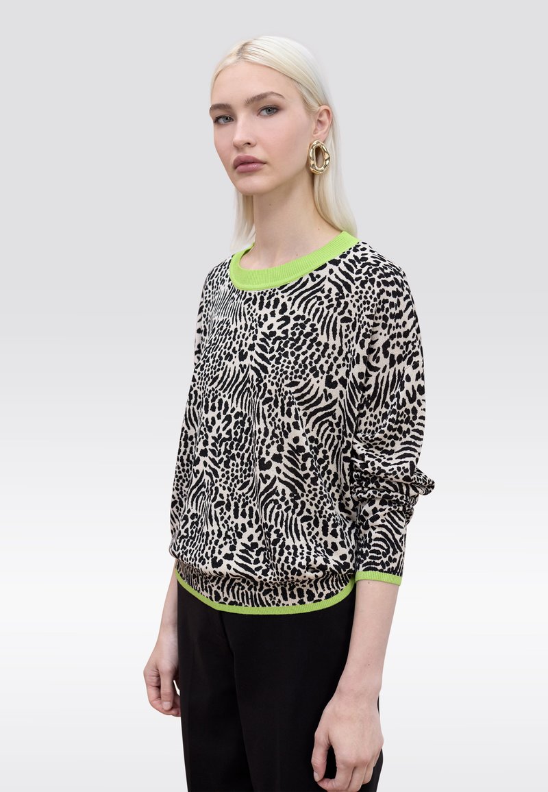 Femme aux cheveux blonds raides portant un pull noir et blanc à motifs animaliers avec des bordures vert néon et un pantalon noir, posant devant un fond uni.