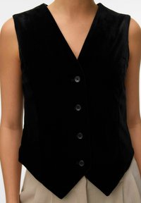 Gilet in velluto nero con scollo a V, senza maniche, e una forma sagomata. Presenta sei bottoni neri sul davanti e un orlo appuntito.